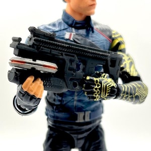 Könnte beinhalten: Eine schwarze und graue Spielzeugpistole mit einem roten und silbernen Aufsatz wird von einer Figur gehalten. Die Figur trägt eine blaue Jacke mit einem weißen Aufnäher und eine schwarze Hose. Der Arm der Figur hat ein gelbes und schwarzes Design.