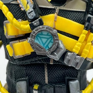 Puede incluir: Primer plano del pecho de una figura con un dispositivo circular metálico con un diseño triangular azul. El atuendo de la figura es negro y amarillo, con correas y detalles amarillos. El dispositivo está sujeto al pecho.