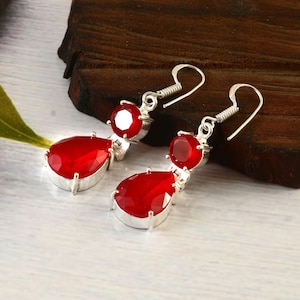 May include: A pair of silver dangle earrings with red gemstone drops. The earrings have a teardrop shape and are accented with smaller round gemstones.