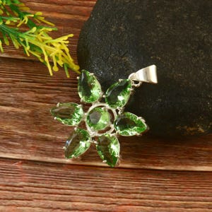 Op de afbeelding: Een zilveren hanger met een bloemmotief en groene edelstenen. De hanger ligt op een houten oppervlak.