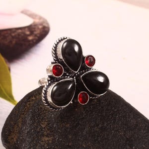 May include: A silver-toned ring featuring a floral design with three large, teardrop-shaped black stones and four smaller, round red stones. The ring is set on a dark, textured surface.