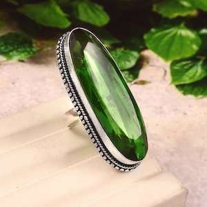 Könnte beinhalten: Ein silberner Ring mit einem großen, ovalen, grünen Edelstein. Der Ring hat einen dekorativen, perlenbesetzten Rand um den Stein.
