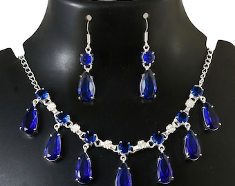 May include: A silver-toned jewelry set featuring a necklace and matching earrings. The necklace has a chain with blue teardrop-shaped pendants and round blue stones. The earrings have a similar design, with a round blue stone and a teardrop pendant.