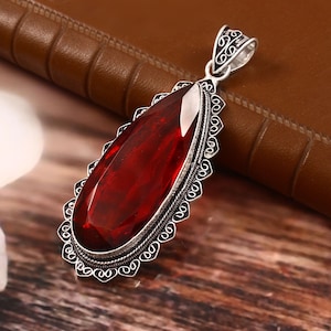 Puede incluir: Un colgante de plata con un diseño adornado presenta una gran piedra preciosa roja en forma de lágrima.