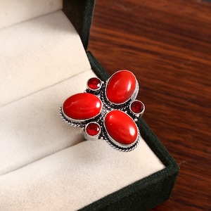 Puede incluir: Un anillo plateado con un grupo de piedras preciosas rojas ovaladas y redondas. El anillo presenta cinco piedras ovaladas grandes y cuatro piedras redondas más pequeñas, todas engastadas en un engaste de metal texturizado y detallado. El anillo se muestra en un joyero verde oscuro.
