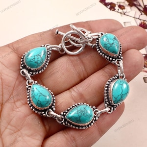 May include: Silver-toned bracelet featuring six turquoise teardrop-shaped stones. Each stone is set in a detailed bezel with a beaded border. The bracelet has a toggle clasp and is displayed on a neutral background.