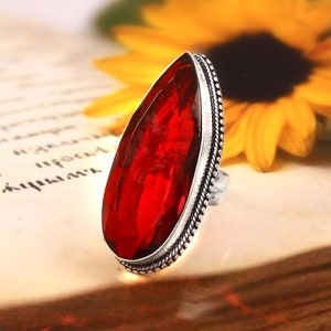 May include: A large, teardrop-shaped red gemstone ring. The faceted stone is set in a silver-colored metal band with a detailed, beaded edge. The ring is displayed on a vintage paper background with a blurred yellow flower.