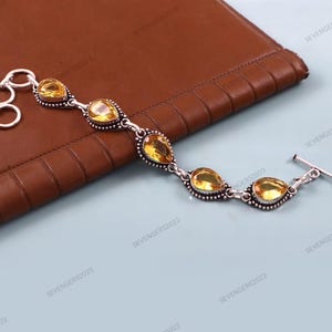 Op de afbeelding: Een zilveren armband met gele, druppelvormige edelstenen. De armband heeft een tuimelsluiting en wordt gepresenteerd op een bruin leren oppervlak. De edelstenen zijn gezet in een decoratieve zilveren zetting.