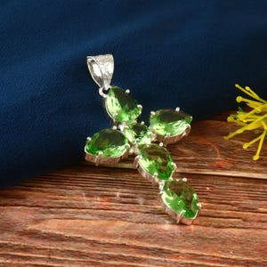 Op de afbeelding: Een zilveren kruis hanger met zes groene edelsteen accenten. De hanger ligt op een houten oppervlak.