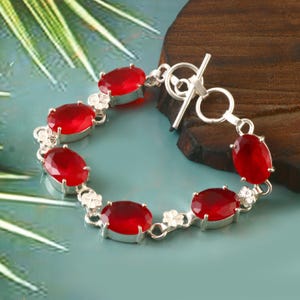 Könnte beinhalten: Ein silbernes Armband mit ovalen, tiefroten Edelsteinen. Das Armband hat einen Knebelverschluss und kleine florale Akzente. Das Armband wird auf einer blau-grünen Oberfläche präsentiert. Die Edelsteine sind ca. 1 cm lang.