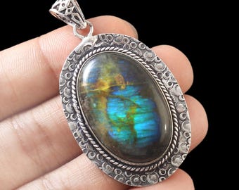 Puede incluir: Pendiente de plata con una gran piedra de labradorita de forma ovalada. La piedra tiene un brillo iridiscente azul, verde y dorado. El pendiente tiene un marco de plata detallado con un diseño texturizado.