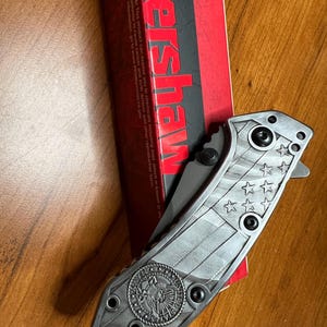 Puede incluir: Una navaja plegable con mango de metal plateado que presenta un patr&oacute;n de bandera estadounidense en relieve y un emblema circular de los Estados Unidos. La navaja est&aacute; colocada junto a su caja de cart&oacute;n roja y negra de la marca Kershaw.