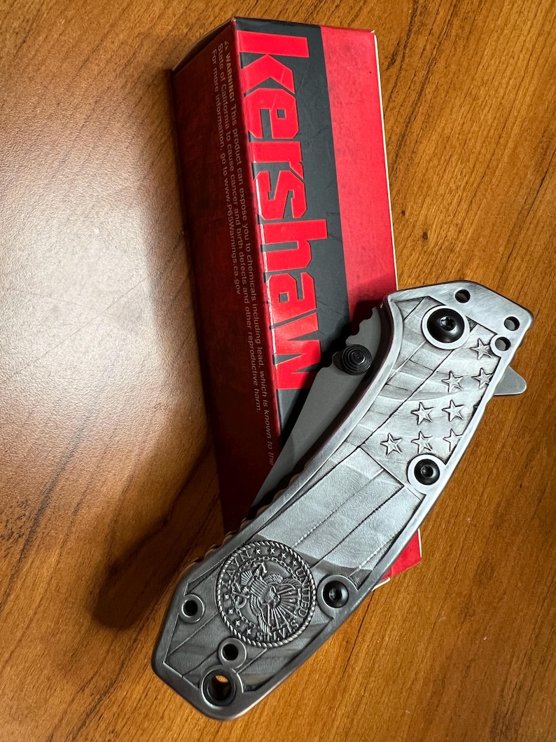 Puede incluir: Una navaja plegable con mango de metal plateado que presenta un patr&oacute;n de bandera estadounidense en relieve y un emblema circular de los Estados Unidos. La navaja est&aacute; colocada junto a su caja de cart&oacute;n roja y negra de la marca Kershaw.