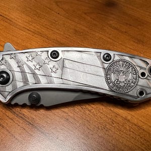 Puede incluir: Una navaja plegable con mango de metal plateado que presenta un dise&ntilde;o en relieve de la bandera estadounidense y un emblema circular de la Marina de los Estados Unidos. El mango incluye tornillos negros y un perno para abrir la hoja.