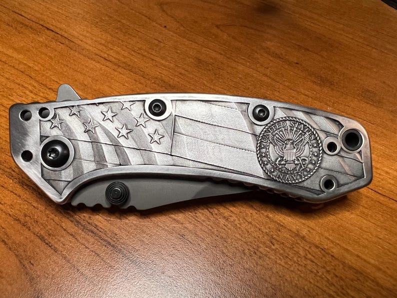 Puede incluir: Una navaja plegable con mango de metal plateado que presenta un dise&ntilde;o en relieve de la bandera estadounidense y un emblema circular de la Marina de los Estados Unidos. El mango incluye tornillos negros y un perno para abrir la hoja.
