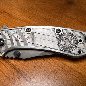 Puede incluir: Una navaja plegable con mango de metal plateado que presenta un patr&oacute;n grabado de la bandera estadounidense y un emblema circular de la Marina de los Estados Unidos. El mango incluye tornillos negros y la hoja parcialmente visible es gris mate.