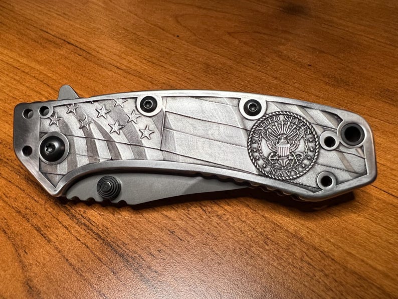 Puede incluir: Una navaja plegable con mango de metal plateado que presenta un patr&oacute;n grabado de la bandera estadounidense y un emblema circular de la Marina de los Estados Unidos. El mango incluye tornillos negros y la hoja parcialmente visible es gris mate.