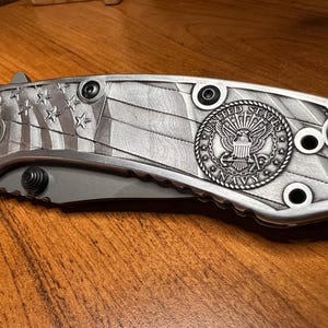 Puede incluir: Una navaja plegable con mango de metal plateado que presenta un patr&oacute;n en relieve de la bandera estadounidense y un emblema circular de la United States Navy. El mango incluye tornillos negros visibles y la hoja permanece cerrada.