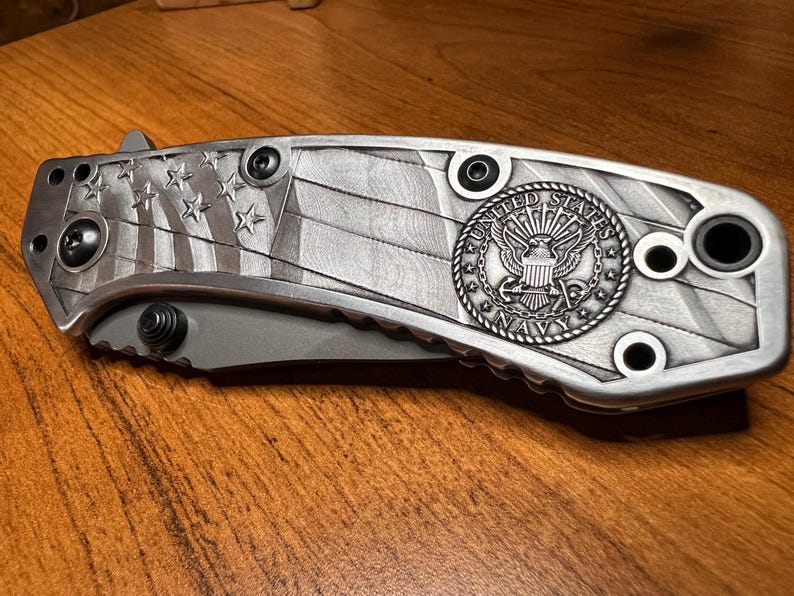 Puede incluir: Una navaja plegable con mango de metal plateado que presenta un patr&oacute;n en relieve de la bandera estadounidense y un emblema circular de la United States Navy. El mango incluye tornillos negros visibles y la hoja permanece cerrada.
