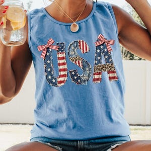 Könnte beinhalten: Ein hellblaues ärmelloses Tanktop mit den Buchstaben USA in einem Patchwork-Design aus amerikanischen Flaggenmustern, Leopardenmuster und Jeans-Texturen, akzentuiert mit zwei kleinen rot-weiß gestreiften Schleifen am U und am A.