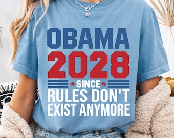 May include: A light blue short-sleeve t-shirt featuring the text OBAMA 2028 SINCE RULES DON'T EXIST ANYMORE in blue, red, and white lettering. The design includes two small red stars and horizontal lines flanking the word SINCE.