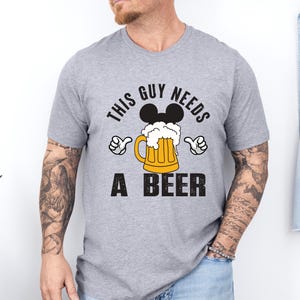 A man wearing a gray t-shirt with a graphic design that says "This guy needs a beer" and features a beer mug with Mickey Mouse ears.