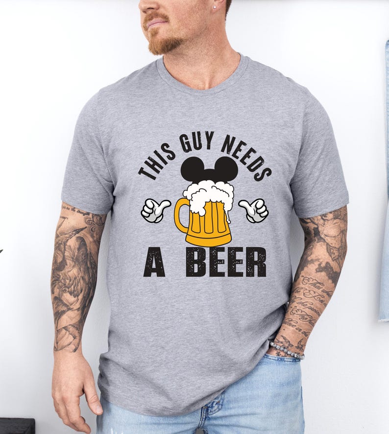 A man wearing a gray t-shirt with a graphic design that says "This guy needs a beer" and features a beer mug with Mickey Mouse ears.