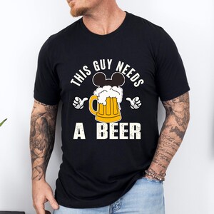 A man wearing a black t-shirt with a graphic design that says "This guy needs a beer" and features a beer mug with Mickey Mouse ears.