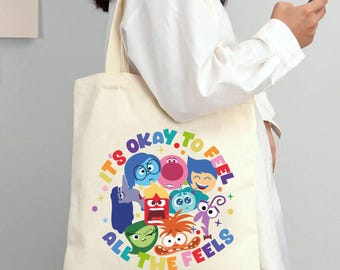 Könnte beinhalten: Cremefarbene Tragetasche mit einem farbenfrohen Grafikdesign von Zeichentrickfiguren und dem Text "IT'S OKAY TO FEEL ALL THE FEELS" in einem Regenbogenbogen. Die Tasche hat lange Henkel und wird von einer Person getragen.