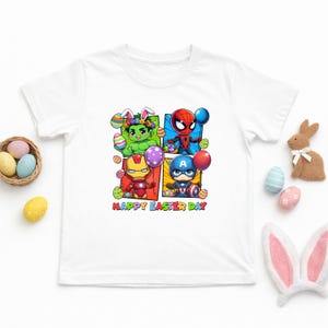 May include: White t-shirt featuring a colorful graphic of Marvel superheroes, including Hulk, Spider-Man, Iron Man, and Captain America, with Easter eggs and the text "HAPPY EASTER DAY". The shirt has a crew neck and short sleeves.
