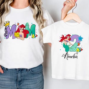 A woman wearing a white t-shirt with the word "MOM" and a smaller t-shirt with the name "Amelia" printed on it, both featuring illustrations of mermaids.
