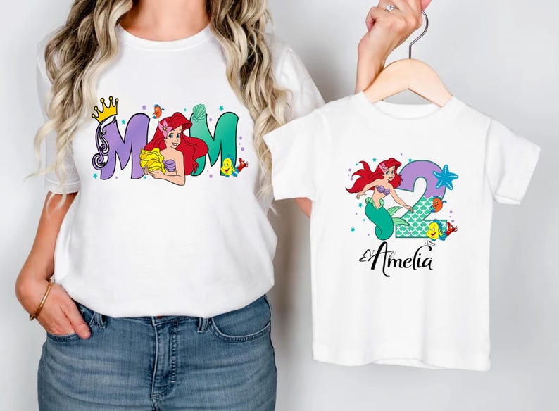 A woman wearing a white t-shirt with the word "MOM" and a smaller t-shirt with the name "Amelia" printed on it, both featuring illustrations of mermaids.