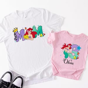 two t-shirts with the same design, one white and one pink, along with a pair of black and white sneakers.
