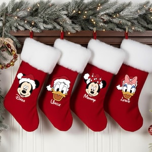 Op de afbeelding: Vier rode kerstsokken met witte imitatiebont manchetten hangen aan een houten schoorsteenmantel. Elke kous heeft een ander Disney-personage: Mickey Mouse, Donald Duck, Minnie Mouse en Daisy Duck, met namen eronder.