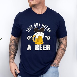A man wearing a navy blue t-shirt with a graphic design that says "This guy needs a beer" and features a beer mug with Mickey Mouse ears.
