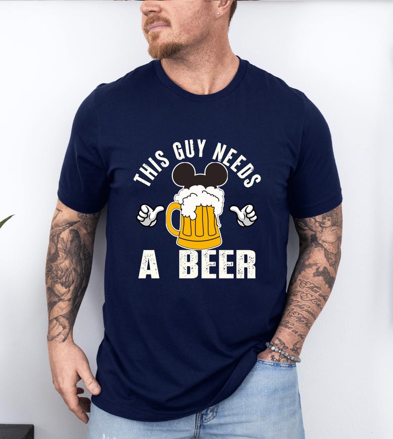 A man wearing a navy blue t-shirt with a graphic design that says "This guy needs a beer" and features a beer mug with Mickey Mouse ears.