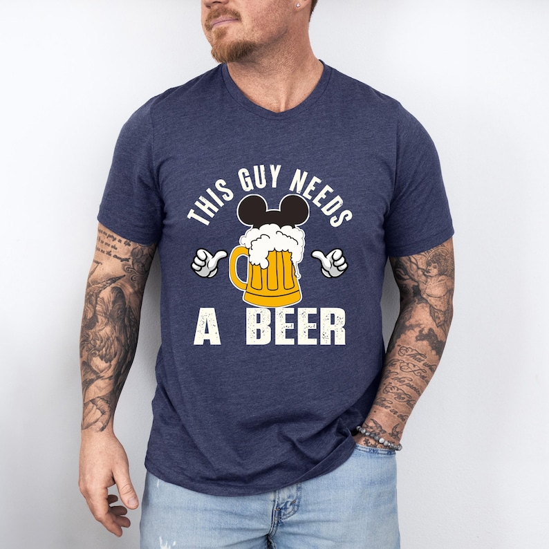 A man wearing a blue t-shirt with a graphic design that says "This guy needs a beer" and features a beer mug with Mickey Mouse ears.