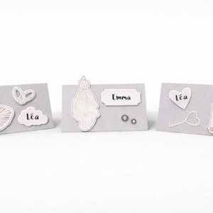 Peut inclure: Trois cartons de table gris avec des éléments décoratifs. L'un présente un flacon de parfum, une fleur et le nom "Léa". Un autre présente une robe et le nom "Emma". Le dernier a un cœur, une clé, un gâteau et le nom "Léa".