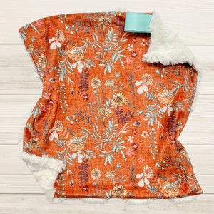 May include: A soft, orange and white floral print baby blanket with a white faux fur border. The blanket has a light blue satin tag attached.