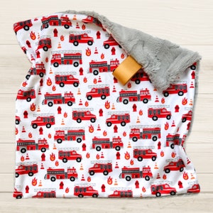 May include: A white baby blanket with a pattern of red fire trucks, fire hydrants, and flames. The blanket has a gray, plush backing and a tan ribbon tag.