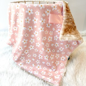 May include: A pink baby blanket with a white daisy print and a brown faux fur backing. The blanket has a pink satin ribbon attached.