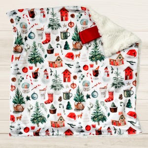 May include: A white baby blanket with a festive Christmas pattern featuring red houses, green trees, ornaments, and candy canes. The blanket has a soft, white fleece backing.