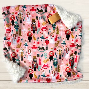 May include: A pink and white baby blanket with a Nutcracker theme. The blanket features a repeating pattern of Nutcracker characters, Christmas trees, and other holiday imagery. The blanket is made of soft, plush fabric and is perfect for cuddling.