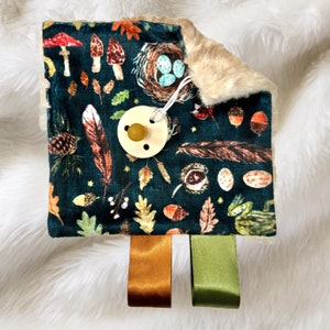 May include: A dark green baby sensory toy with a woodland theme. The toy features a variety of textures, including soft fur, smooth wood, and crinkly fabric. It also has a pacifier clip and three satin ribbons in orange, green, and brown.