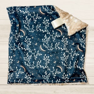 May include: A soft, plush throw blanket with a dark blue background and a white floral pattern featuring crescent moons. The blanket has a faux fur trim.