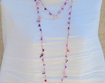 Puede incluir: Un collar largo de varias capas con cuentas y piedras rosas y moradas. El collar está ensartado en un fino cordón rosa y se exhibe en un maniquí blanco. Las cuentas y piedras varían en tamaño y forma.