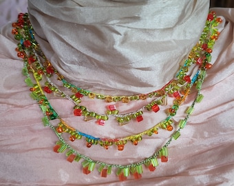 May include: A multi-strand beaded necklace with a variety of colorful beads. The necklace features green, orange, red, and yellow beads, with a delicate, dangling design. The necklace is displayed on a light pink fabric.
