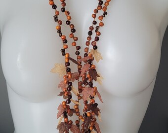 May include: A multi-strand necklace with wooden beads in shades of brown and copper. The necklace features leaf and grape cluster pendants. The necklace is displayed on a white mannequin.