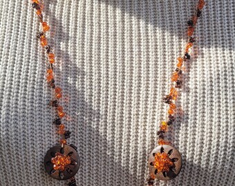 May include: A handcrafted necklace featuring a beaded chain in shades of orange and brown. The necklace has four dark wooden pendants with intricate cut-out designs and orange bead clusters. The necklace is displayed against a cream-colored sweater.
