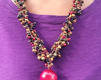 May include: A multi-strand necklace with a large, round, red pendant. The necklace is made of small, colorful beads in shades of red, black, brown, and yellow. The person wearing the necklace is wearing a purple shirt.